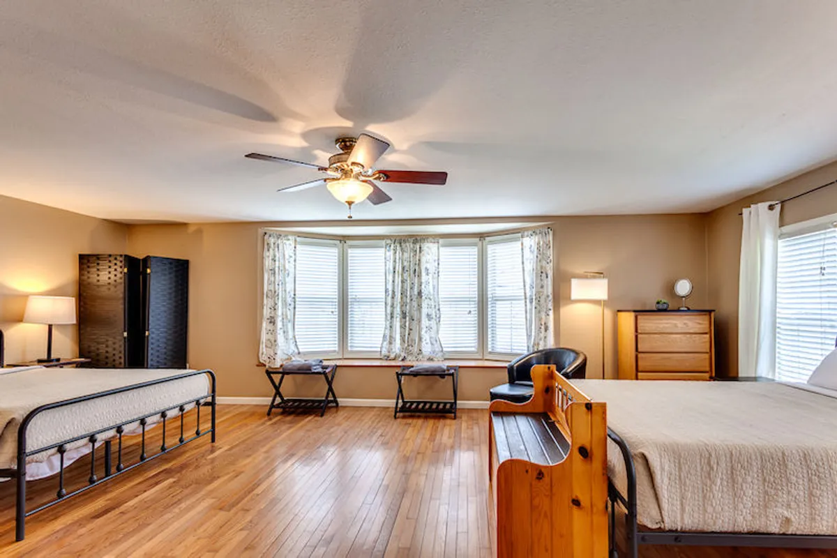 A spacious bedroom showcasing two queen beds, wooden flooring, a bay window for abundant daylight, and a ceiling fan overhead. The setup is comfortable and ideal for families or friends with plenty of room to relax and unwind.