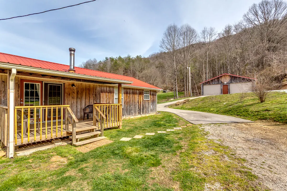 The image presents a welcoming view of a wooden porch with a red metal roof, stone pathway, and lush greenery surrounding the property. Outbuildings and nature trails are visible, suggesting plenty of space for exploration and outdoor activities.