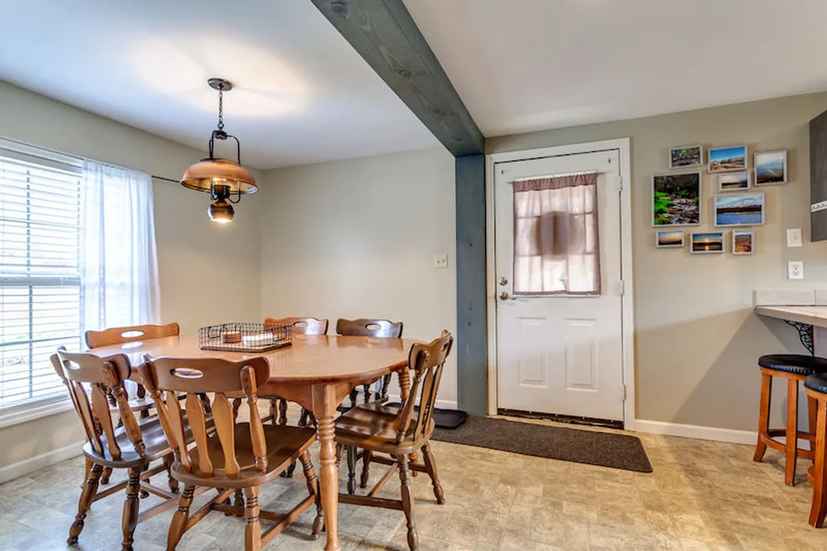 An inviting dining area centered around a wooden table set for six, beneath a stylish hanging light fixture. The space feels warm and communal, with family photos on the wall and a clear view of the open doorway, suggesting connection to other living areas.