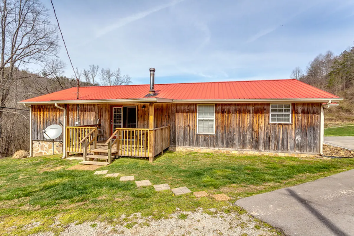 A cozy wooden cabin with a red metal roof and a welcoming front porch. The surrounding natural landscape features lush greenery, and the porch offers a comfortable spot to enjoy the outdoors, whether for morning coffee or sunset views.