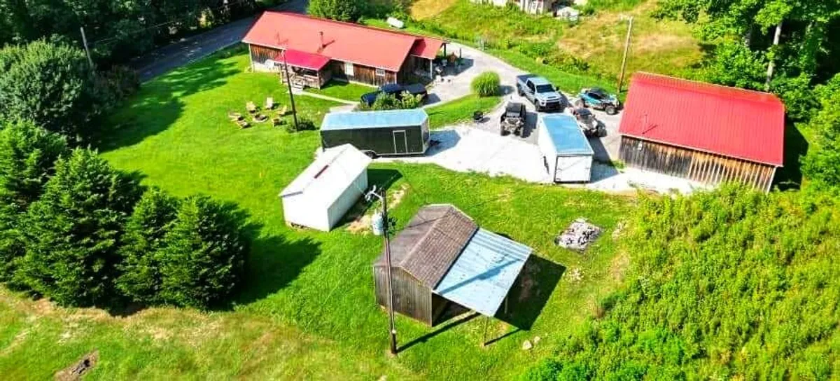 A broad view of red-roofed buildings set amid a green lawn, with various outbuildings and vehicles, creates a rustic, peaceful atmosphere. The space hints at opportunities for outdoor gatherings, barbecues, and exploring the surrounding property.