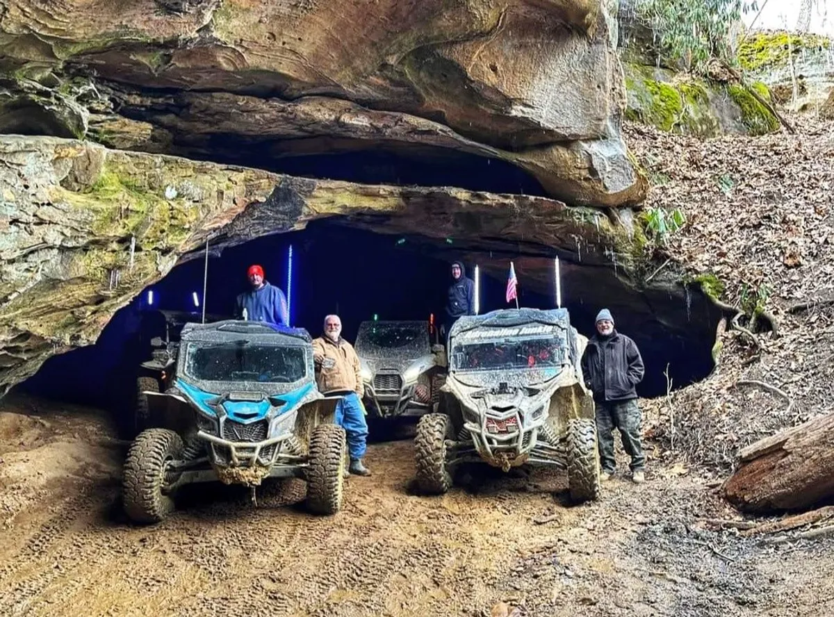 A group of people stands near ATVs beside a rocky cave, ready for off-road adventure. The scene captures excitement and camaraderie, with the wild, wooded landscape hinting at the nearby trails and possibilities for exploration.