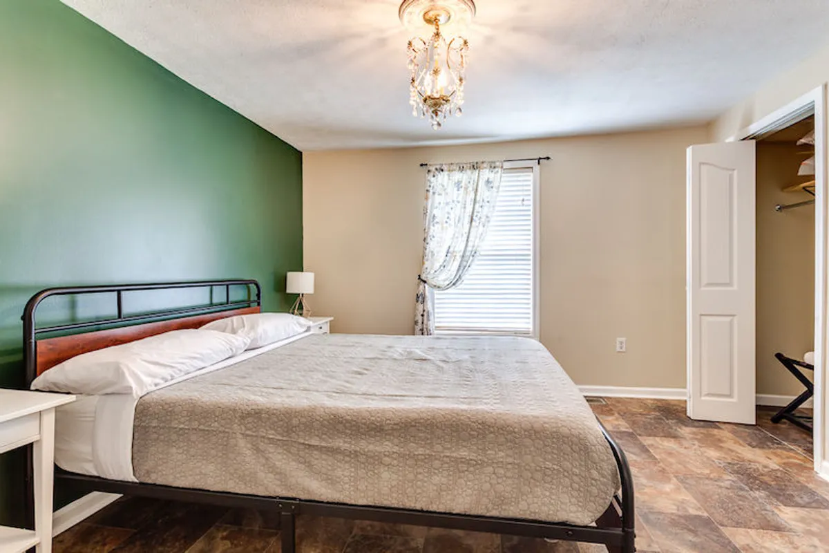 A calming bedroom featuring a comfortable bed with rich linens, a green accent wall, a window draped with soft curtains, and a decorative chandelier overhead. Natural light streams in, creating a serene atmosphere ideal for rest and relaxation.