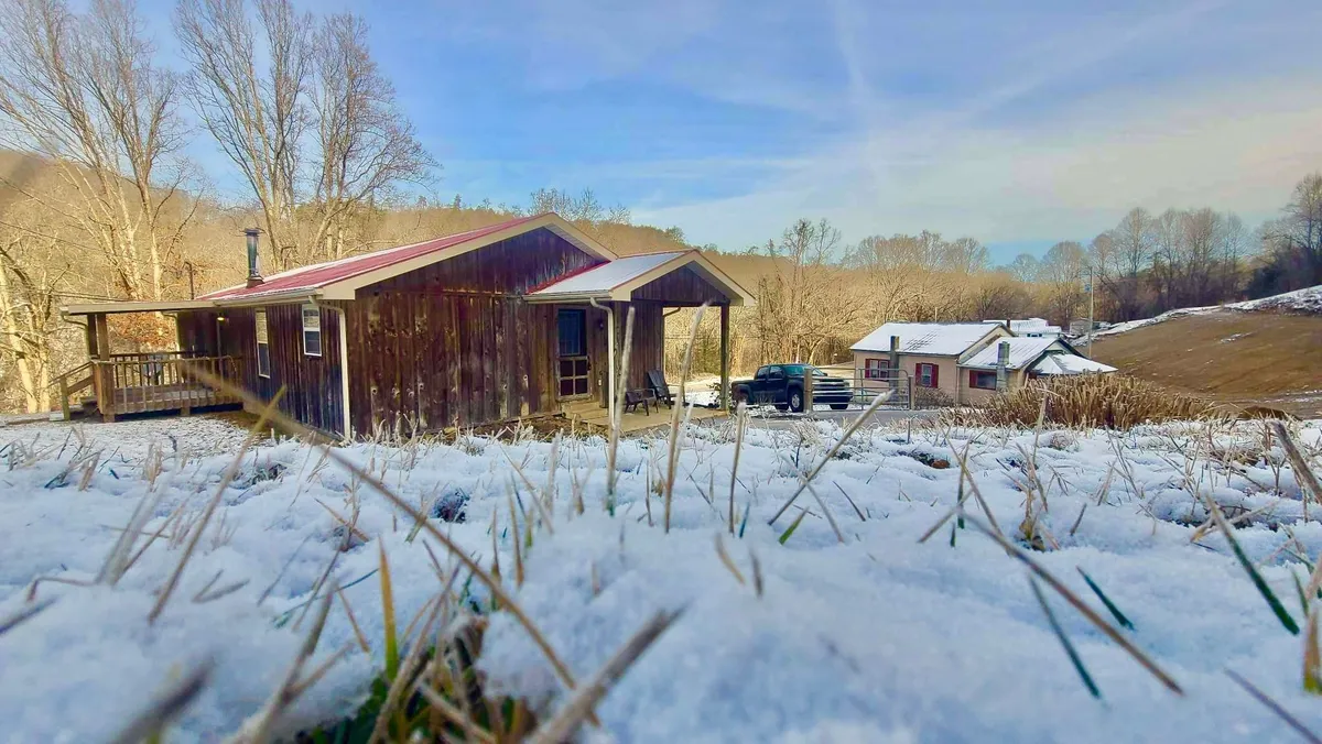 A wooden cabin nestled among snow-covered ground and trees, with distant hills, captures a peaceful winter scene. The porch and yard suggest family-friendly activities like watching snowfall or gathering by the firepit, even in colder months.