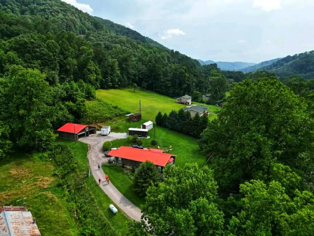 The image showcases a charming red-roofed home set amidst lush greenery, with a winding driveway leading up to the house. The backdrop features rolling, distant mountains under a serene sky, suggesting sunset or golden hour. The inviting landscape hints at outdoor gatherings, relaxation, and connection with nature.