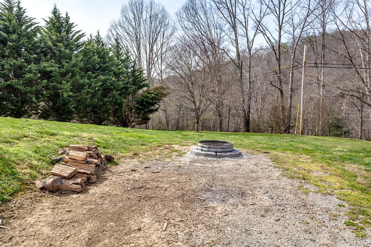 A tranquil outdoor fire pit surrounded by trees, with a ready stack of firewood nearby. The natural clearing offers a peaceful atmosphere perfect for evening gatherings. The area is set up for campfire activities such as storytelling and roasting marshmallows.